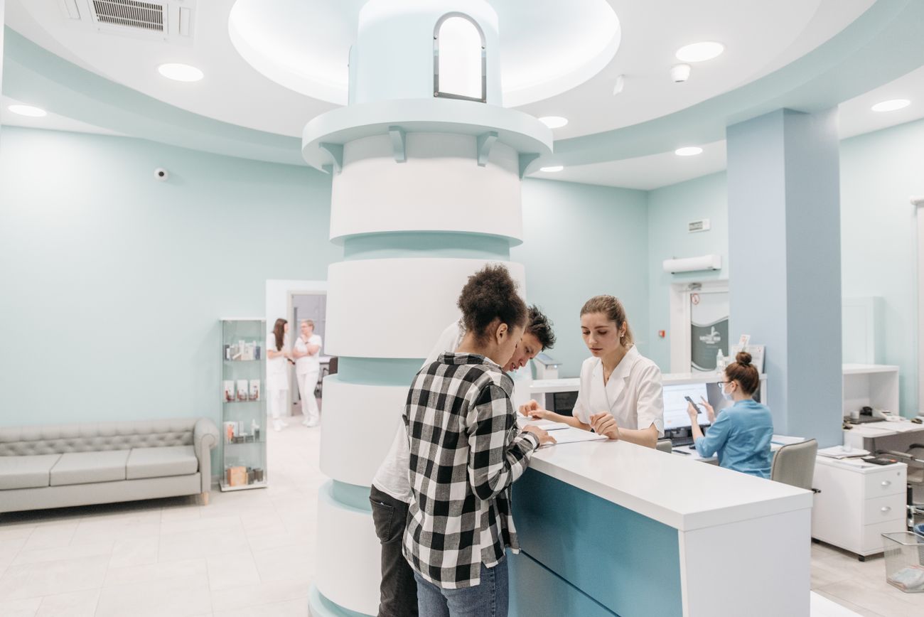 Two people at a clinic reception counter, seen from across the hall