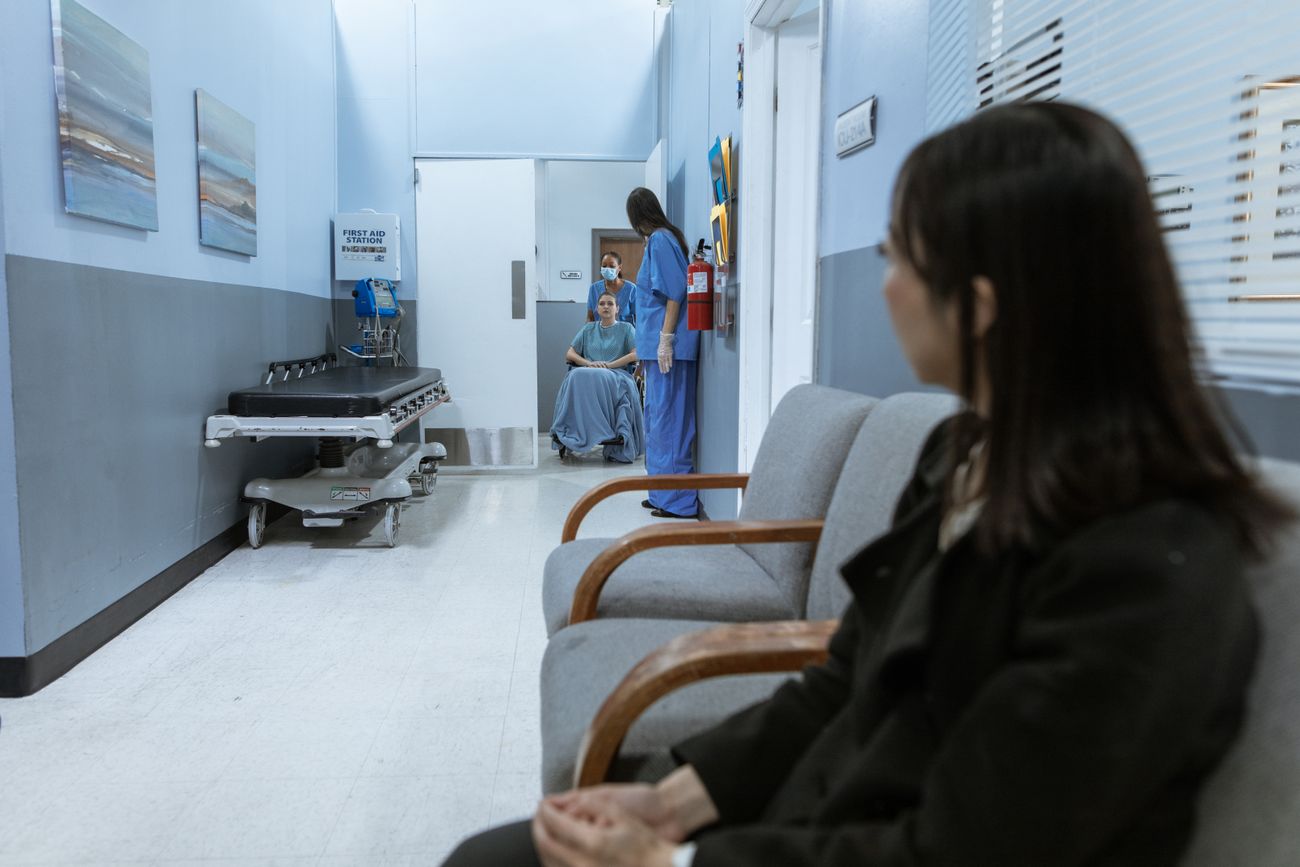 People waiting on chairs in a hospital corridor while staff move a patient